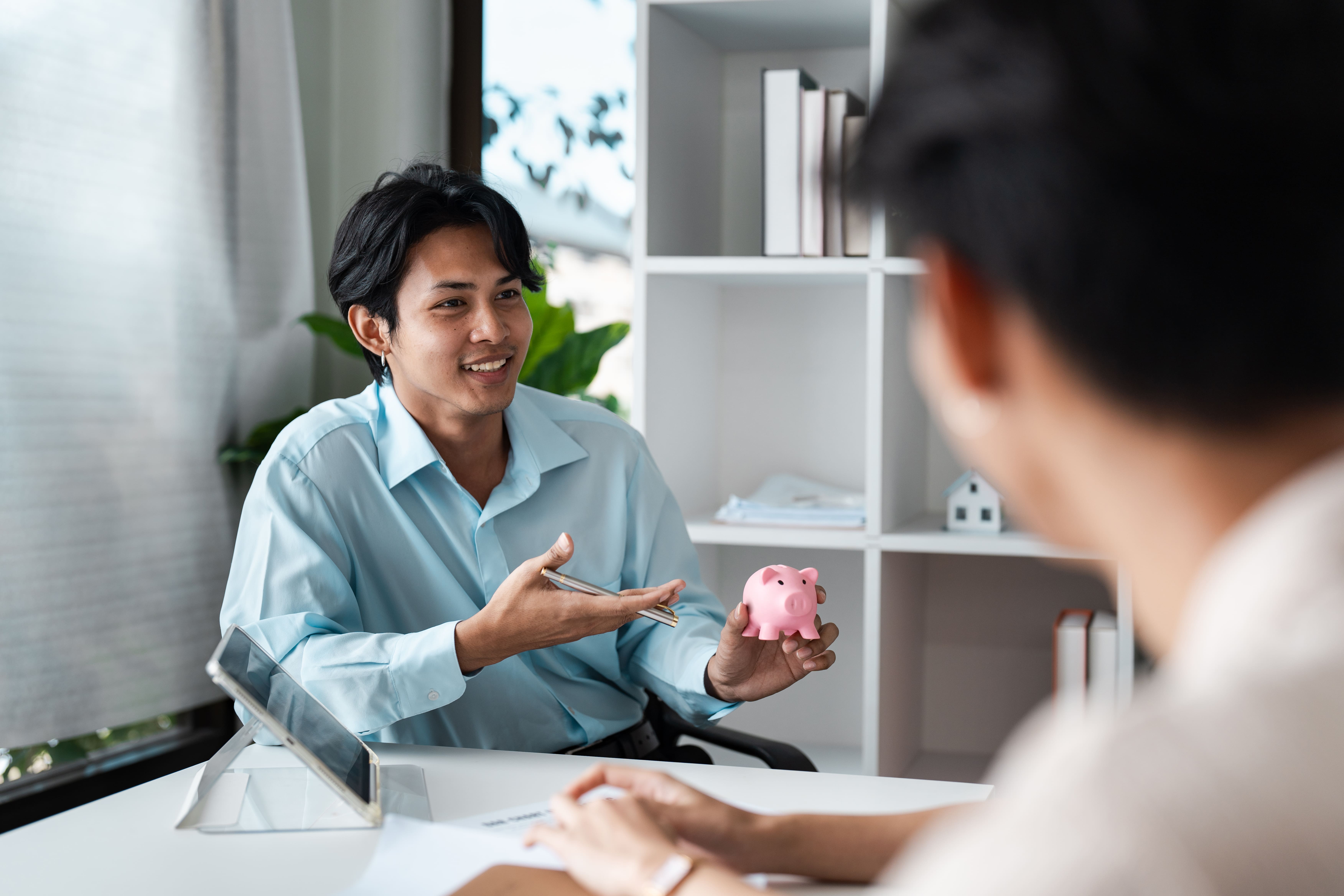 Smiling financial advisor explaining savings strategy while holding a pink piggy bank during client meeting.