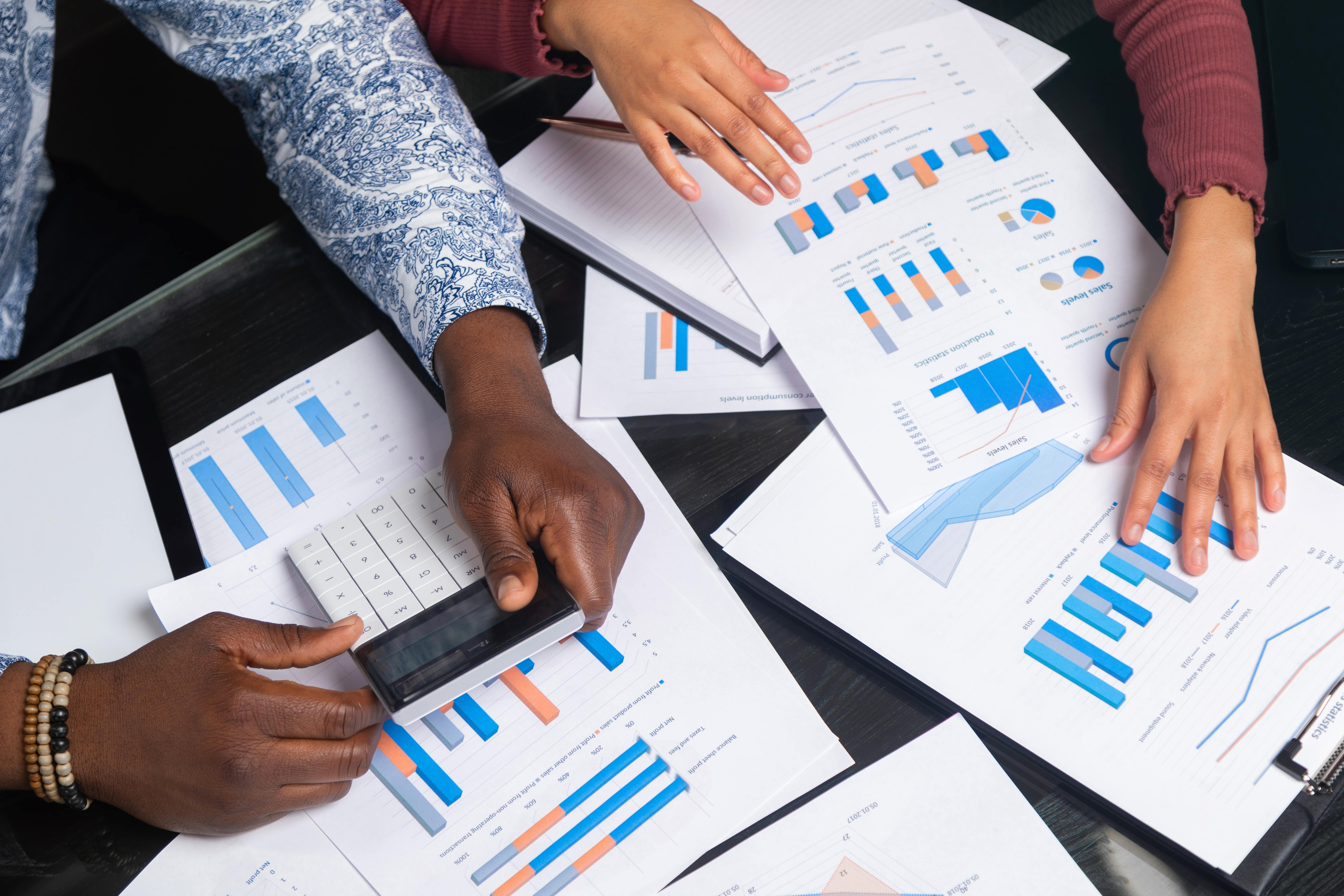 Hands of two people reviewing financial charts and data reports with a calculator on a desk.